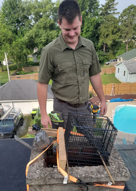 A man in a green shirt and tan pants holds a large turtle with both hands. He stands on a paved driveway with a residential area in the background. A black SUV is parked nearby, and the surroundings include trees and grassy lawns under a sunny sky.